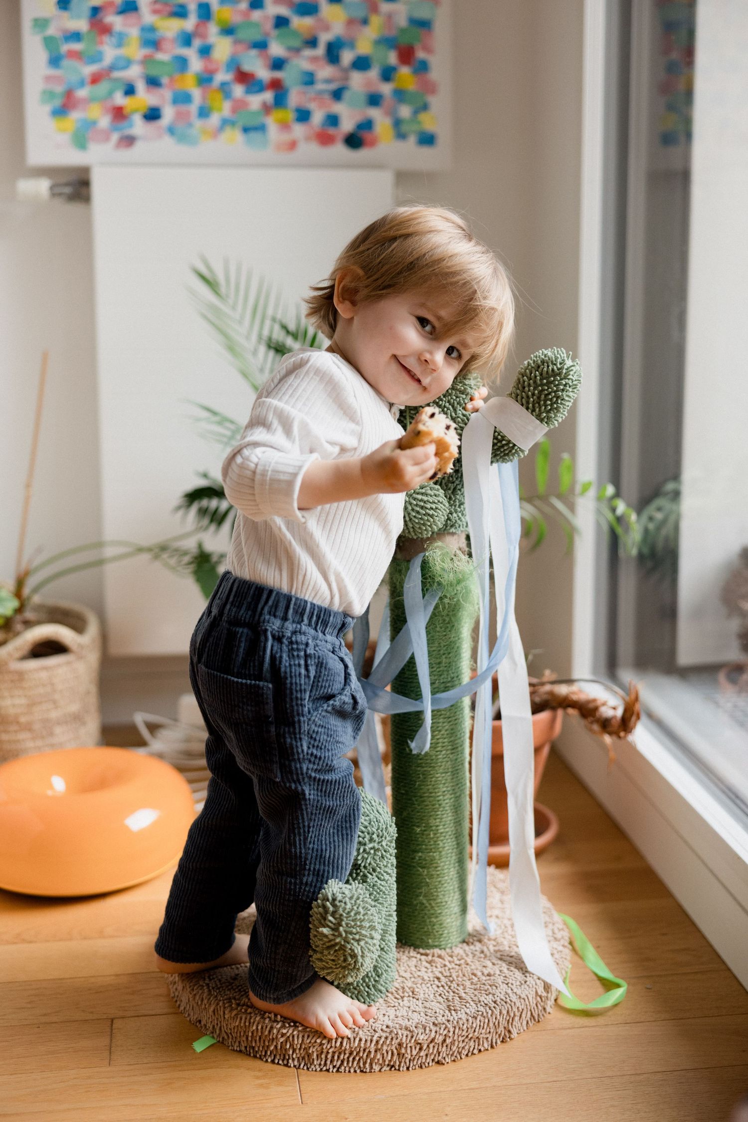 Smiling toddler boy playing with a green cactus cat tree toy indoors, wearing white shirt and jeans.