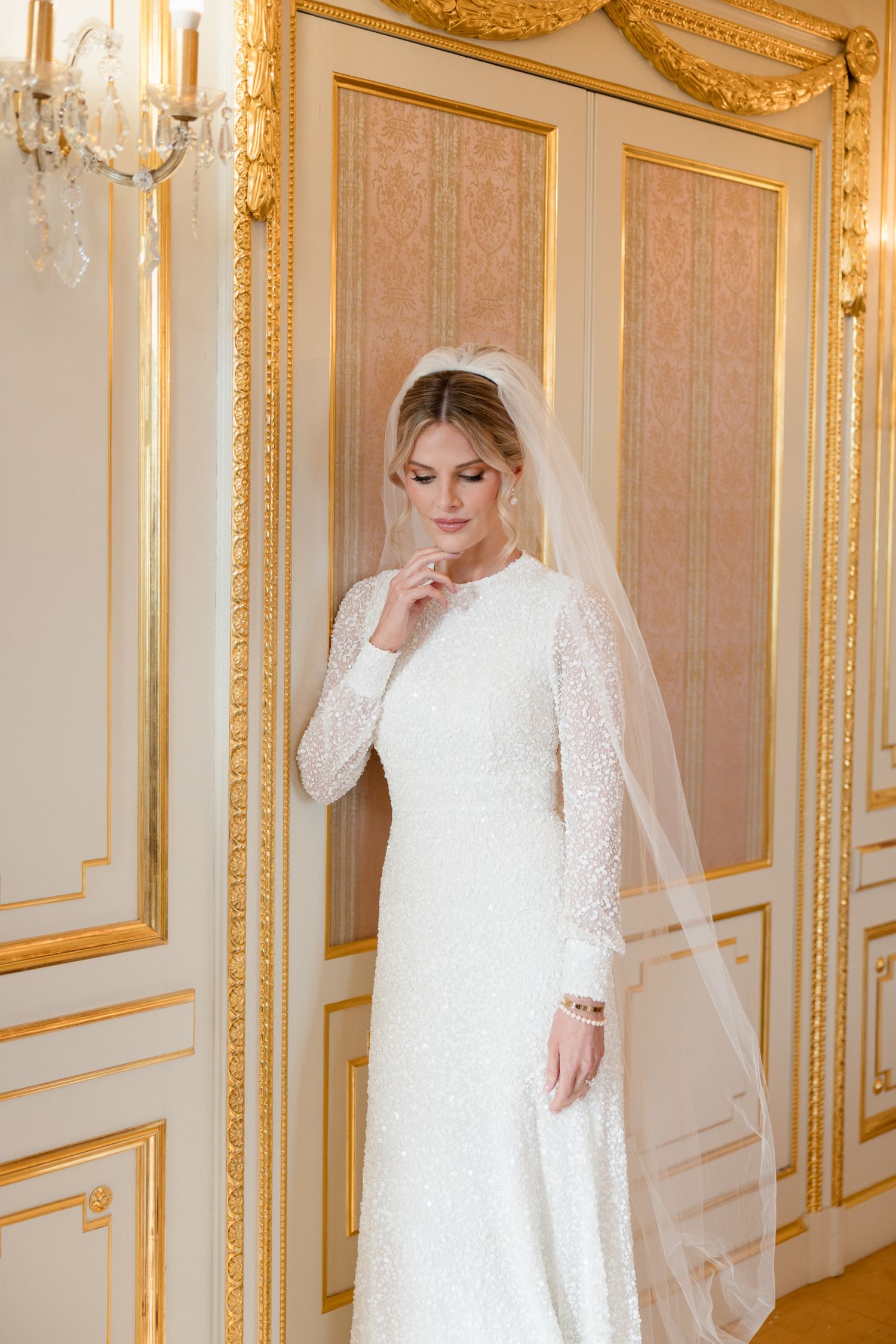 Bride in elegant white lace long-sleeve wedding gown poses in opulent room with gilded gold trim and crystal chandelier.