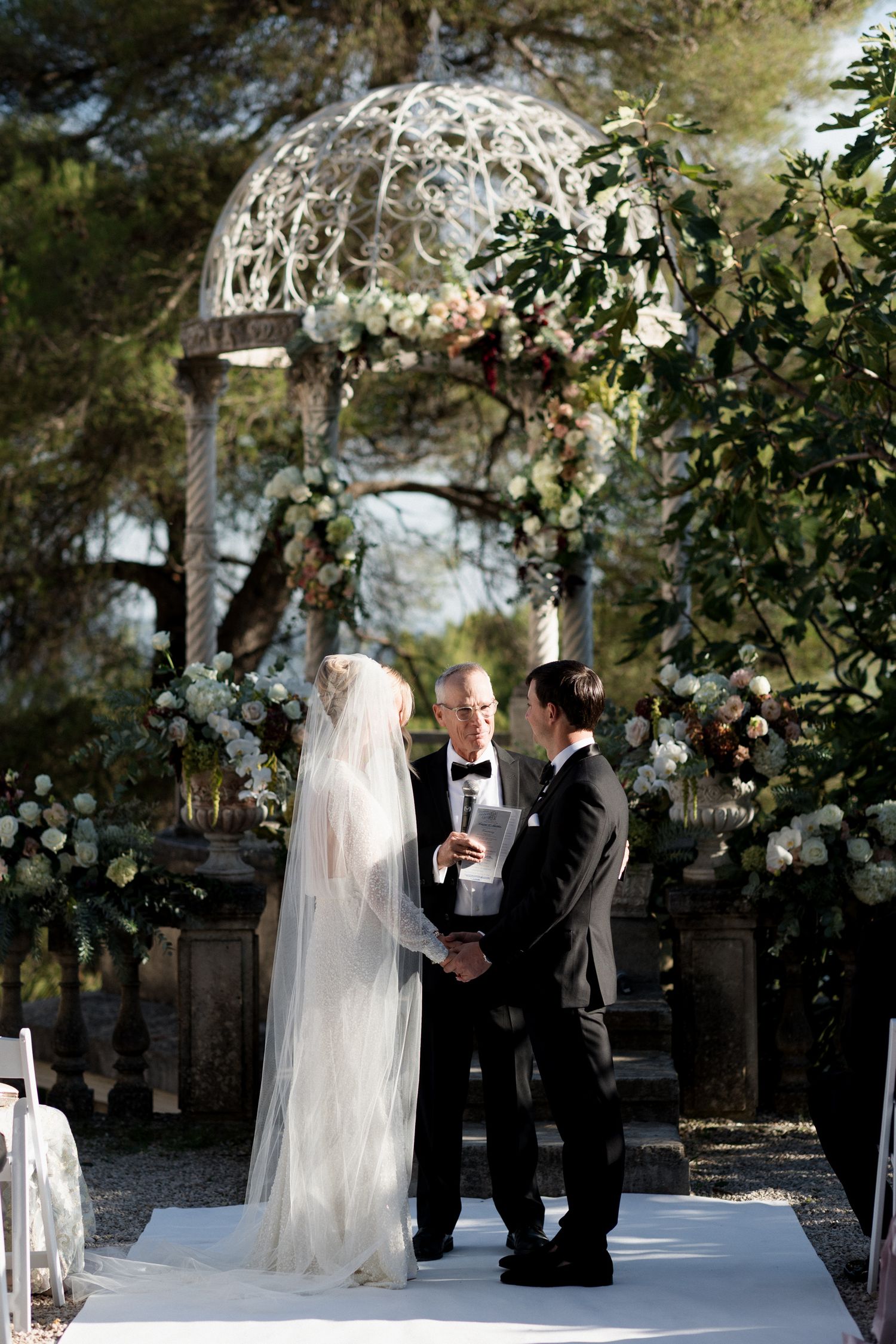 Wedding ceremony taking place under a decorated outdoor gazebo with greenery and floral arrangements.