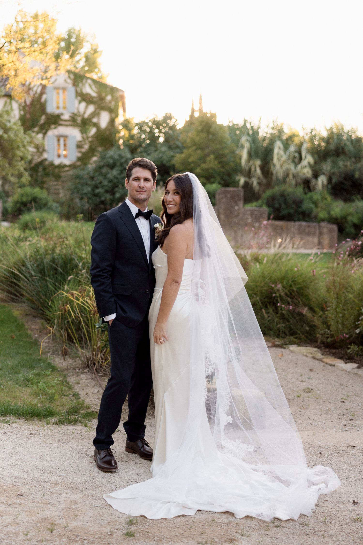 A romantic wedding couple poses together on a garden path with a flowing white veil trailing behind them. wedding photographer france