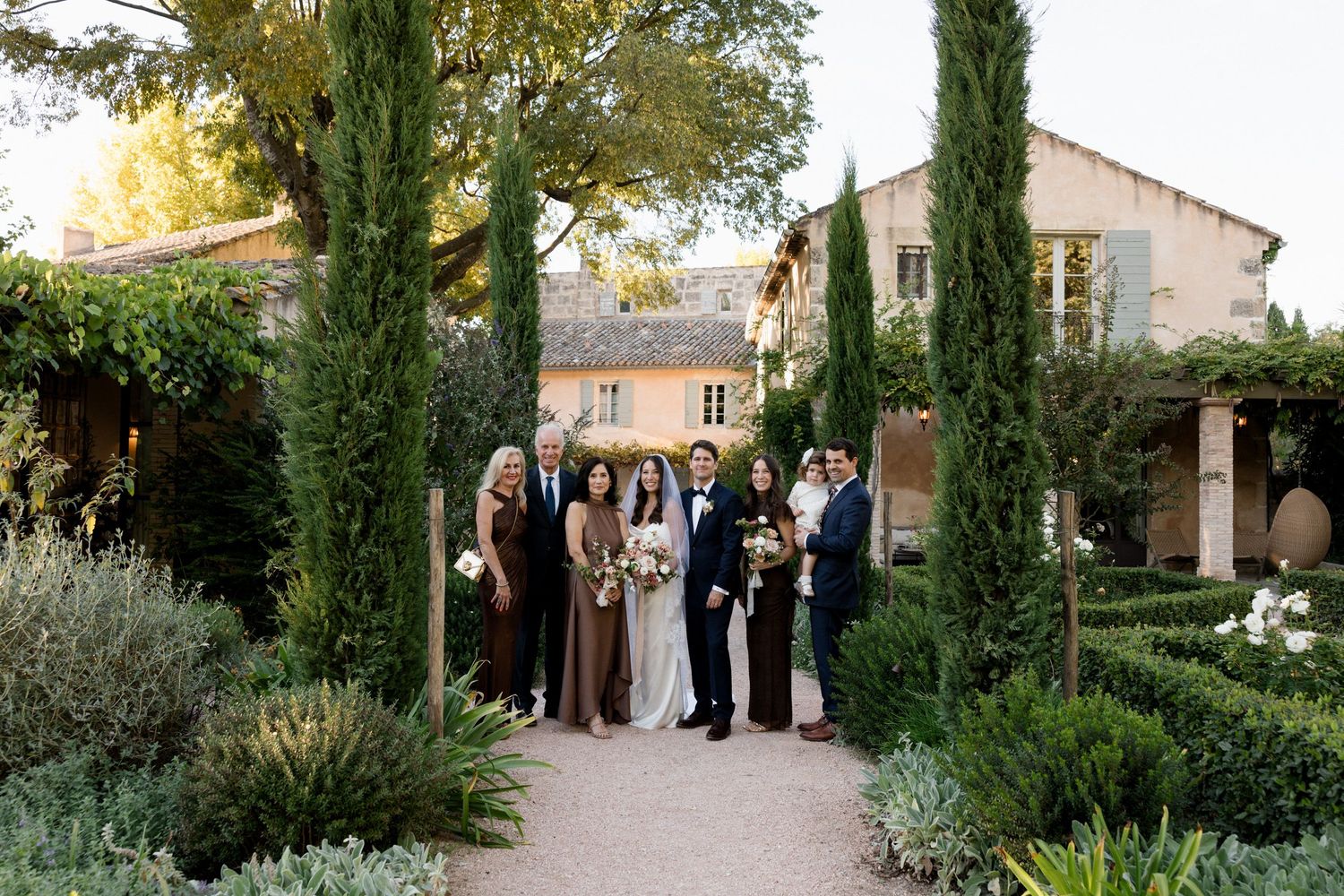 A wedding party gathers in a lush Mediterranean garden with tall cypress trees and stone architecture.
