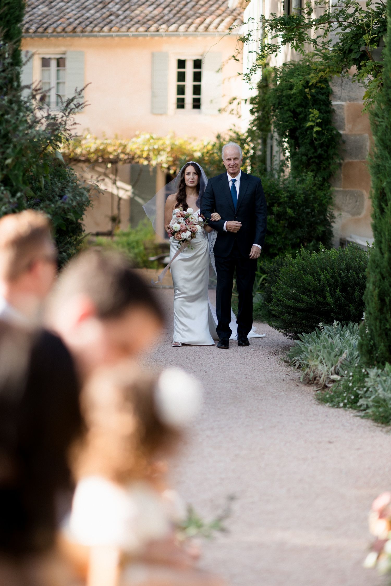 A bride and her escort walk down a sunlit garden path during a wedding ceremony procession.