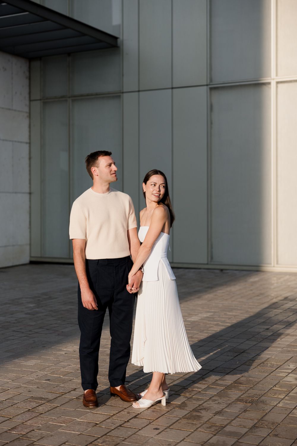 A couple stands together in an industrial space wearing contrasting white dress and dark pants against a concrete backdrop. Seance couple To