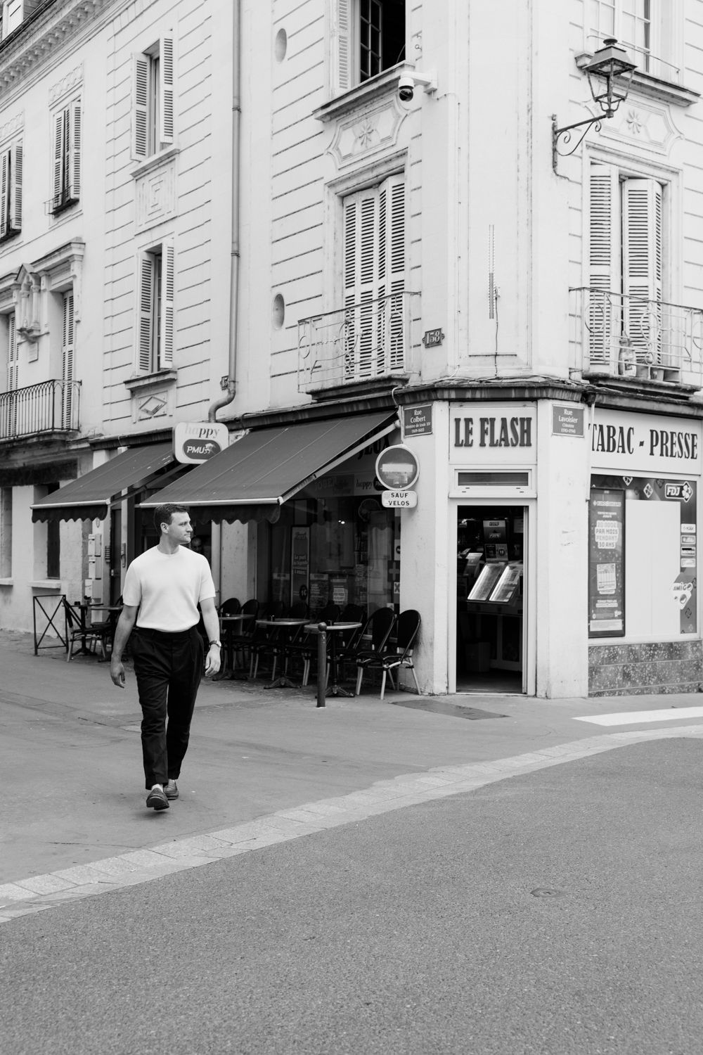 People walking along a quiet French city street lined with classic white buildings and storefronts in black and white.