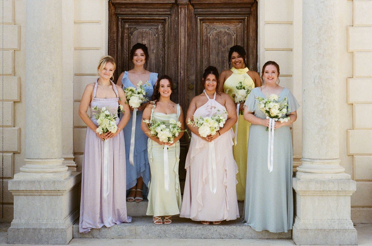 Wedding party group poses in pastel dresses against stone pillars of a classic building exterior. Analog wedding Château des 3 Fontaines