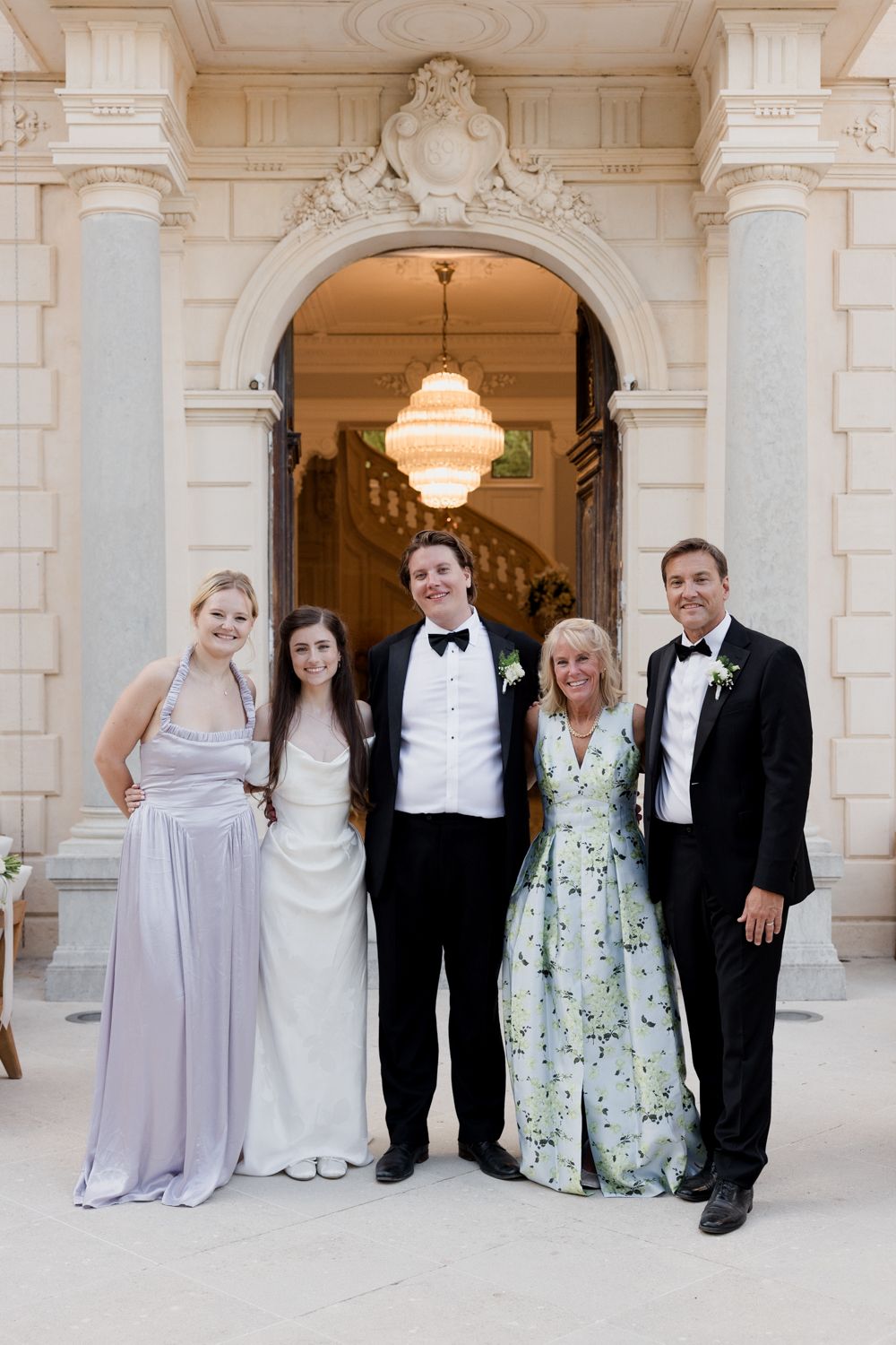Wedding party poses together in formal attire against elegant white marble columns in classical venue.