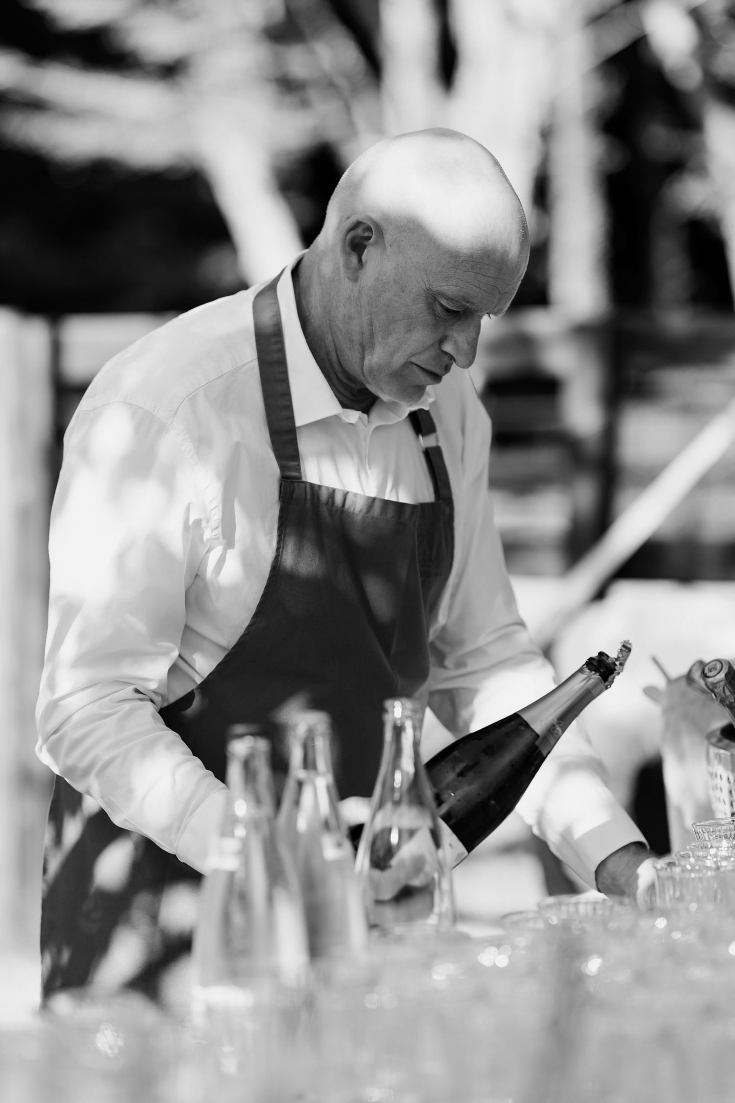 Black and white panoramic image shows bartender in white shirt preparing drinks behind a bar with multiple glasses.