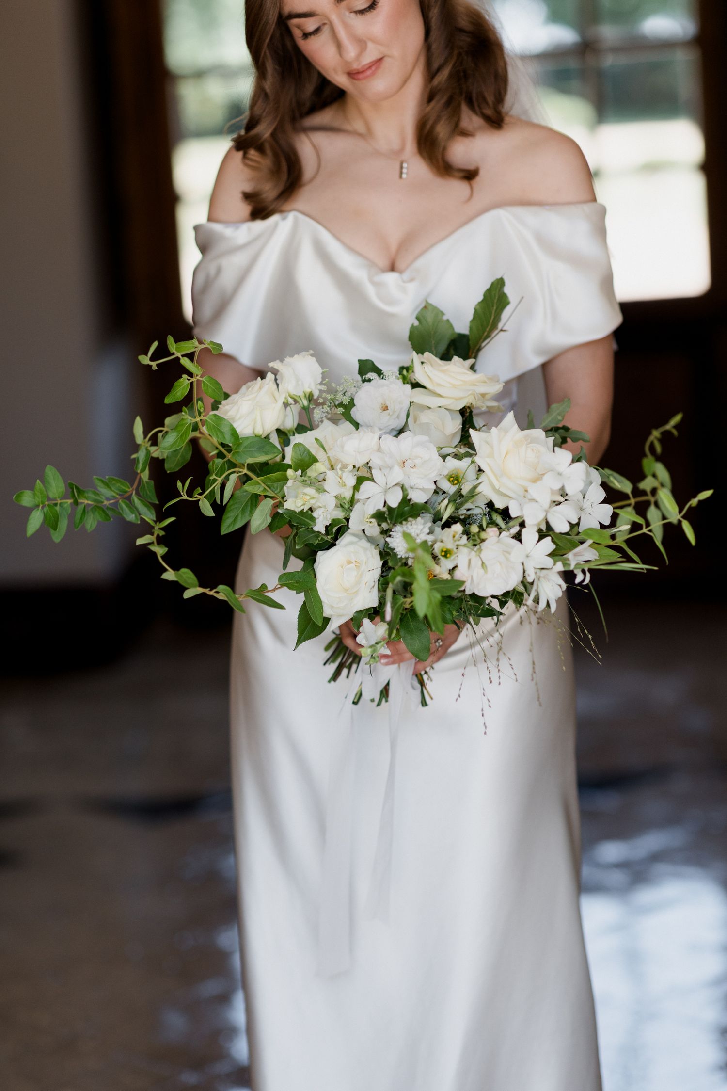 A bride in a classic white off-shoulder wedding dress holds an elegant bouquet of white flowers and greenery.
