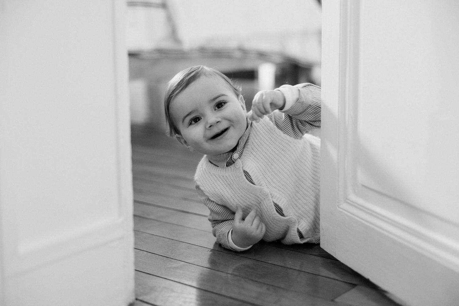 Black and white photo of a happy toddler crawling on hardwood floor, smiling and pointing toward camera in a doorway.