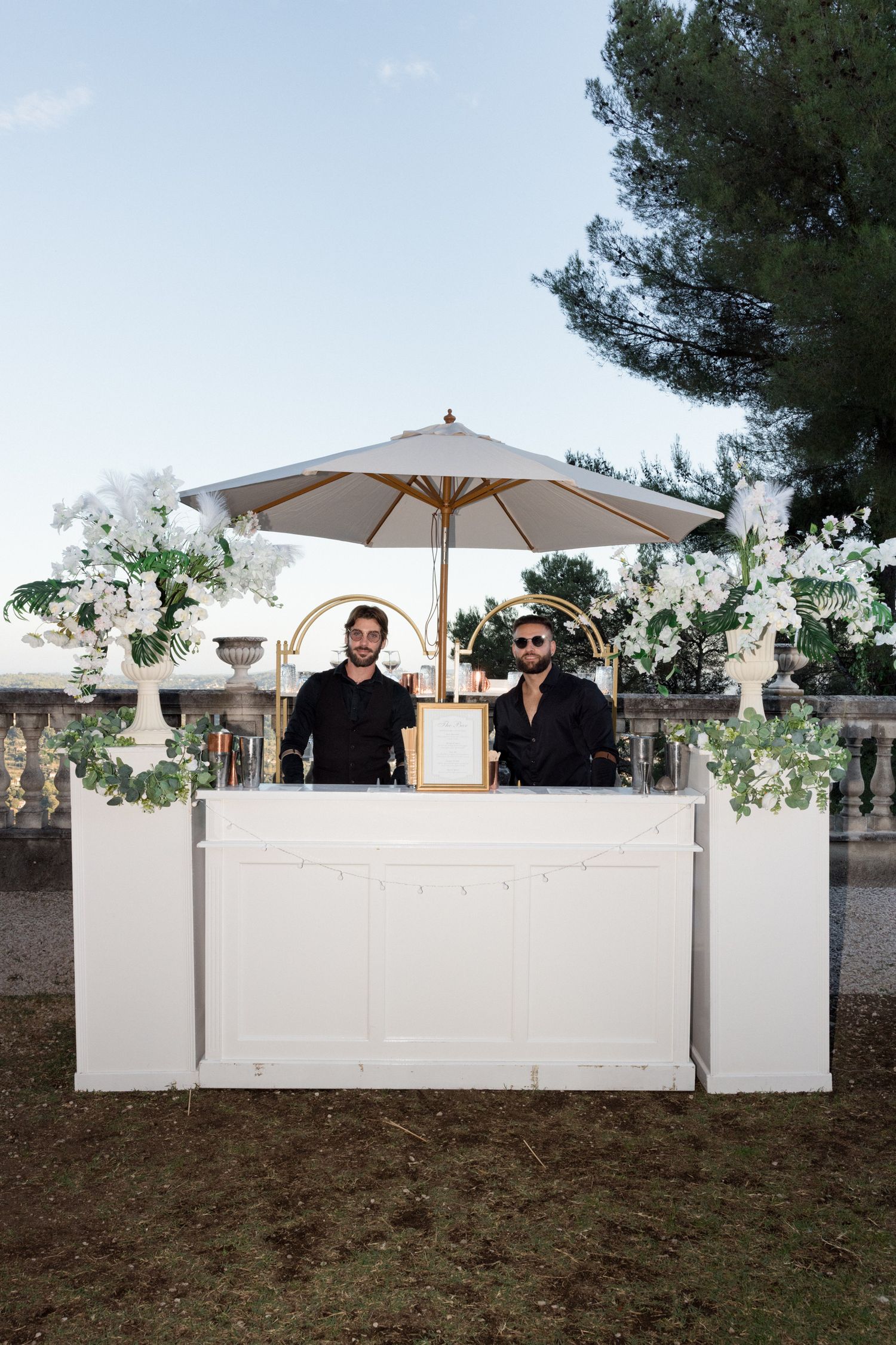 An elegant outdoor bar setup with a white counter, beige umbrella and floral arrangements at an event.