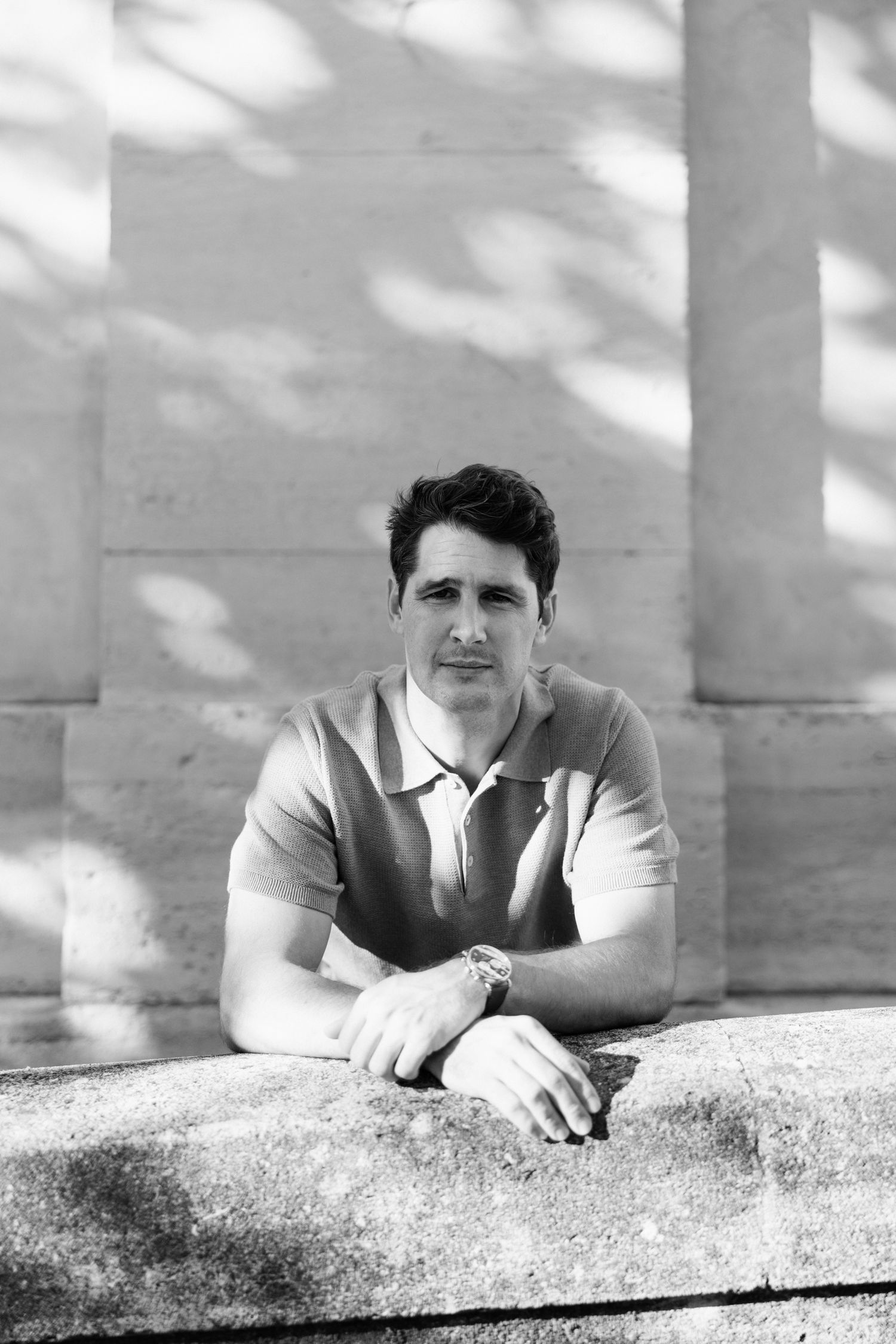 A person in a white polo shirt leans against a stone wall in a black and white artistic portrait composition.
