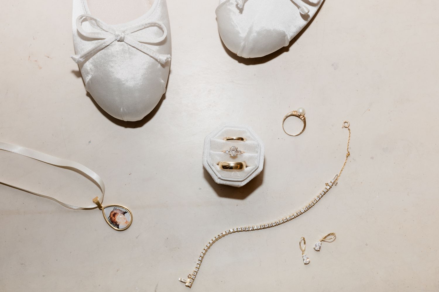 Bridal accessories including white ballet flats, delicate gold jewelry, and a ring box arranged on white background.