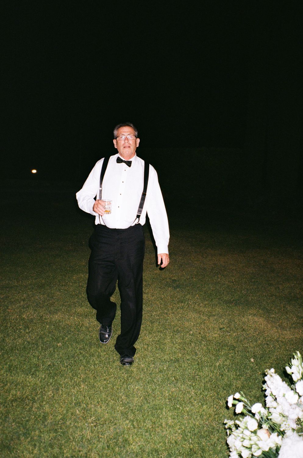 A person in formal attire with suspenders runs through a grass field at night past white flowers.