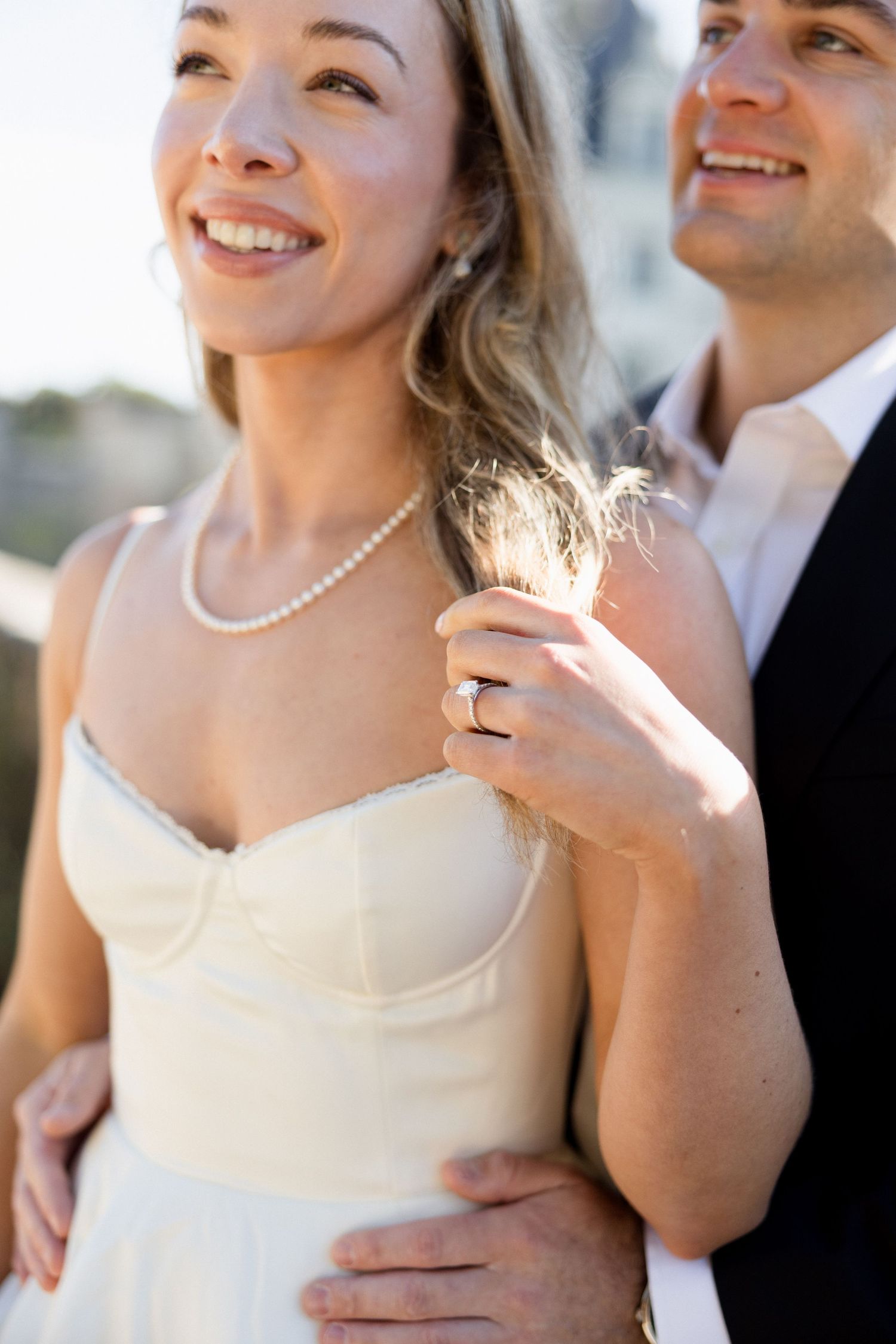 Smiling bride in white dress with pearl necklace and groom embracing outdoors in golden light.