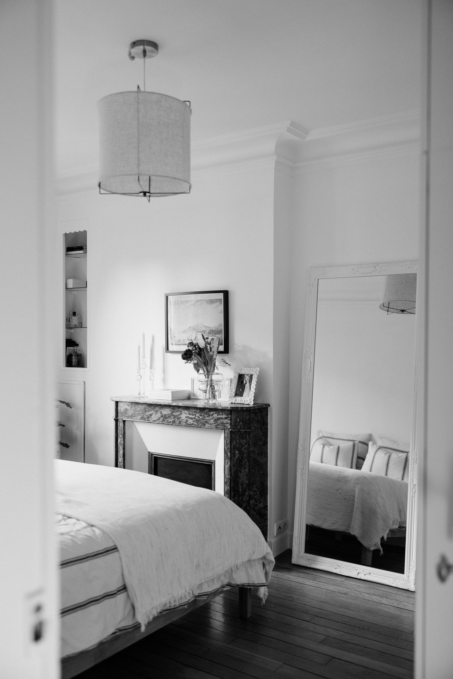 Elegant black and white bedroom with pendant light, marble fireplace, tall mirror, and neatly made bed on dark hardwood floors.