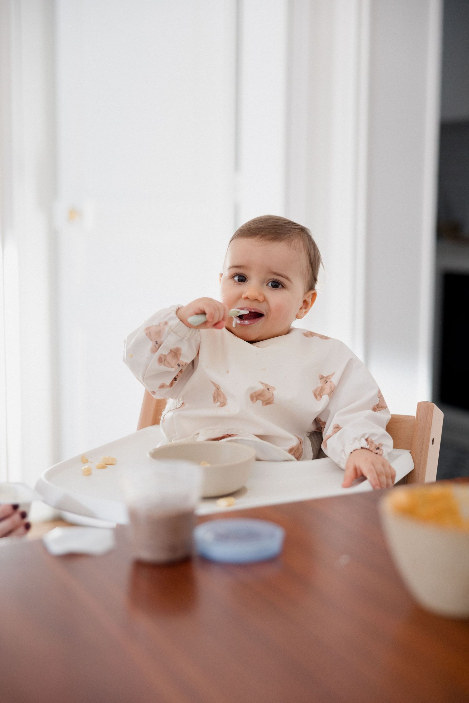 Cute baby in a patterned bib self-feeding with a spoon in a high chair at a wooden dining table.