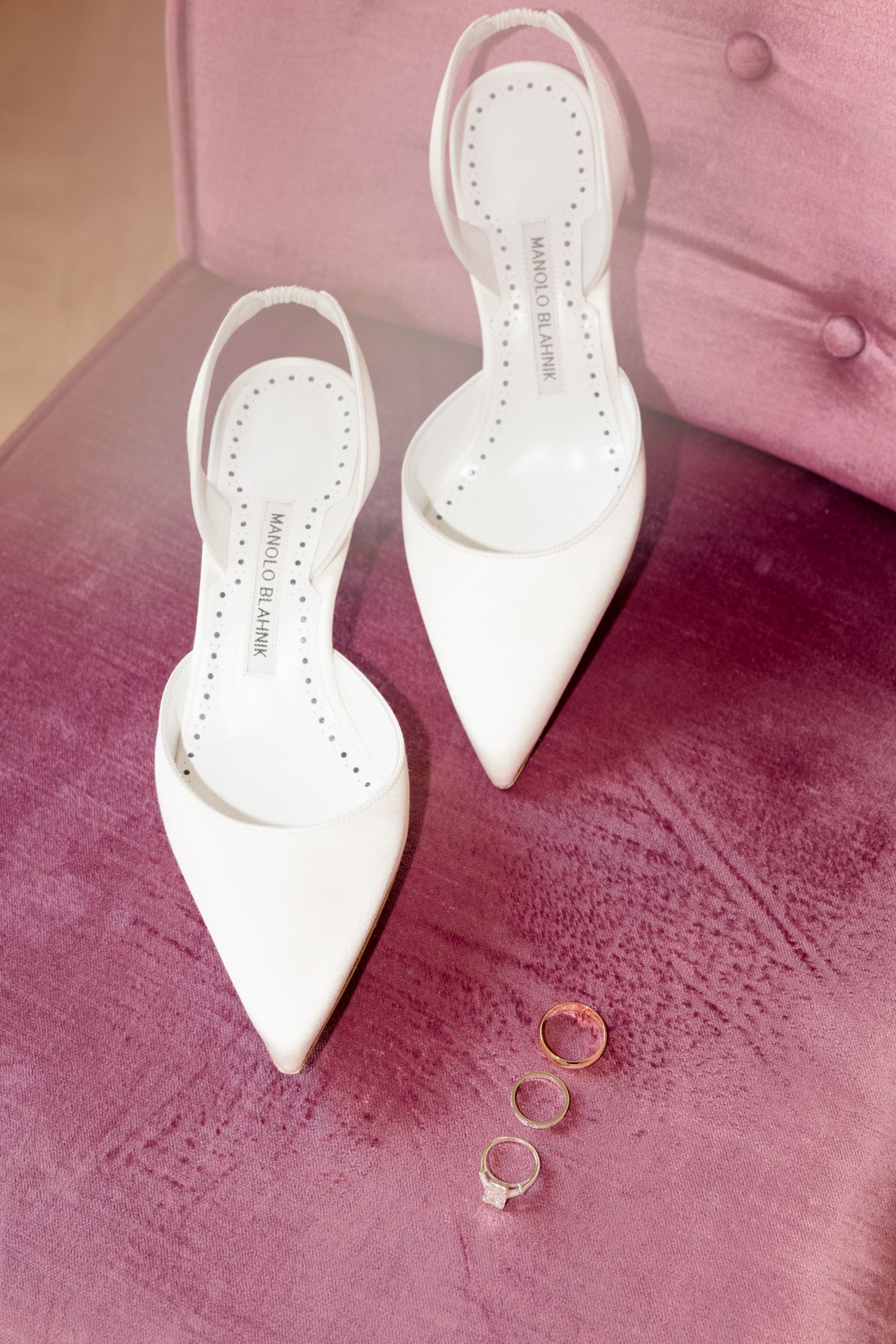 White pointed stiletto heels and gold wedding rings arranged on a pink velvet surface in a romantic composition. destination wedding france