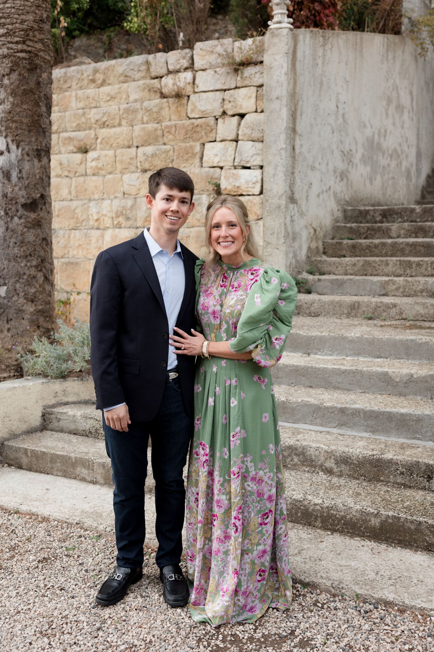 A couple stands together on stone steps in front of a historic wall, with the woman wearing a green floral dress.
