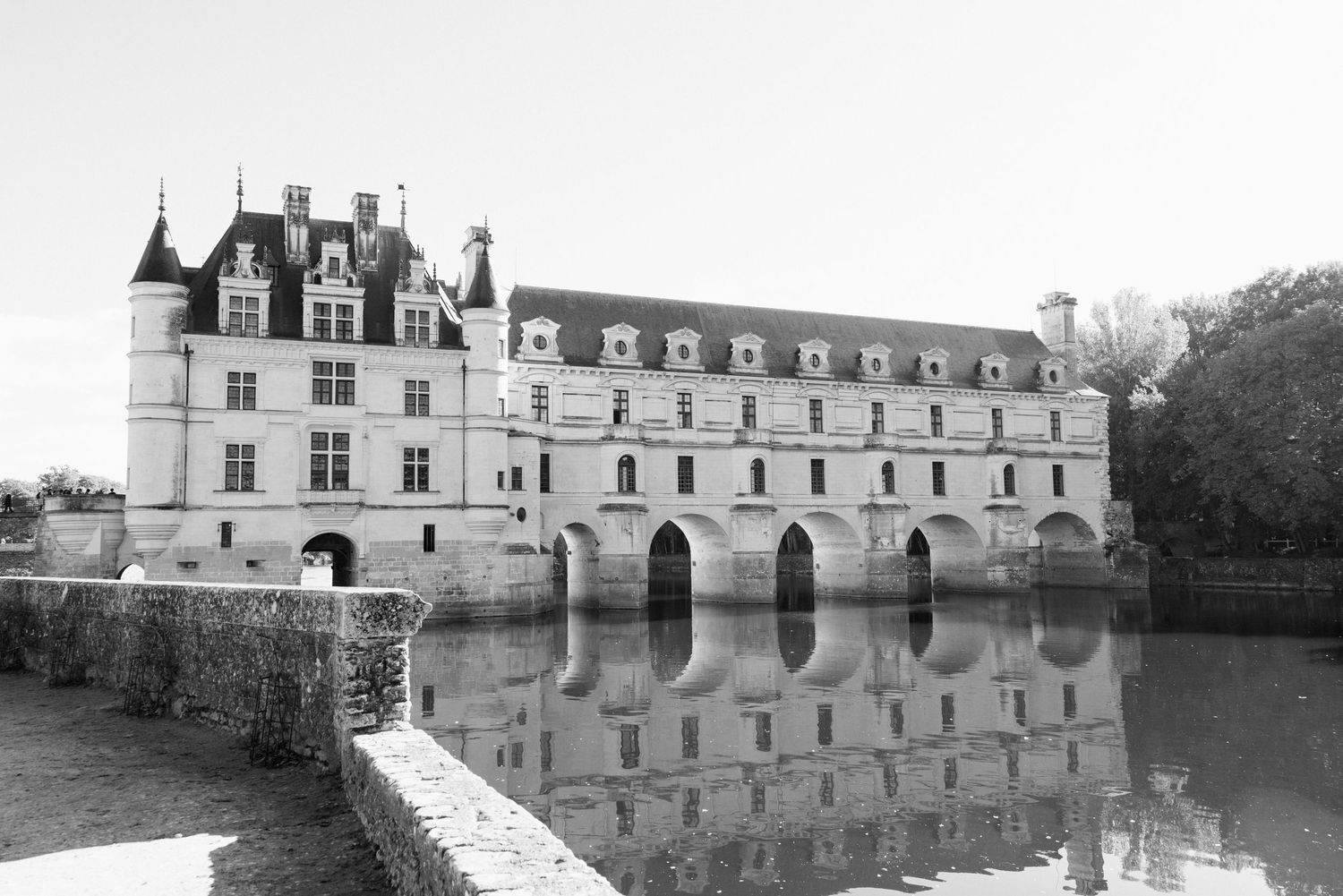Black and white photo of Chateau de Chenonceau reflecting in calm river waters on a clear day.