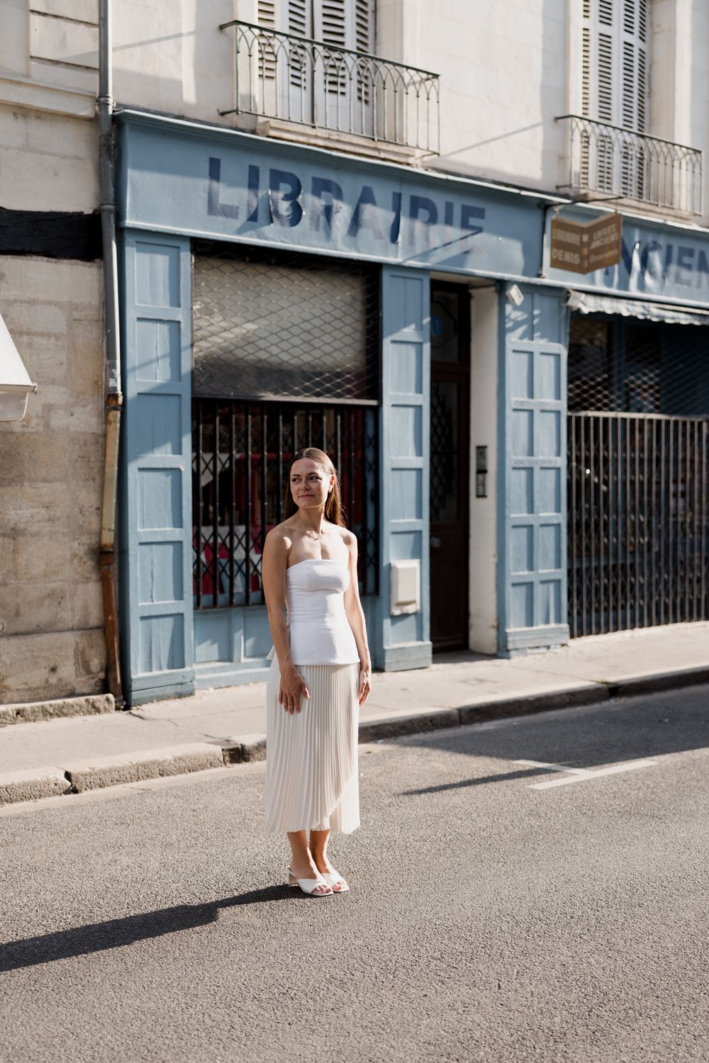 A woman in a white sleeveless dress stands on a quiet city street with light blue storefronts and shuttered windows.