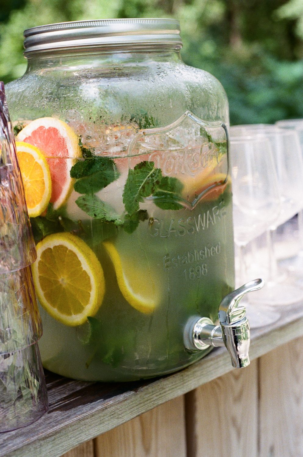 Glass beverage dispensers filled with citrus infused lemonade sit on white wooden railing in bright sunlight outdoors.
