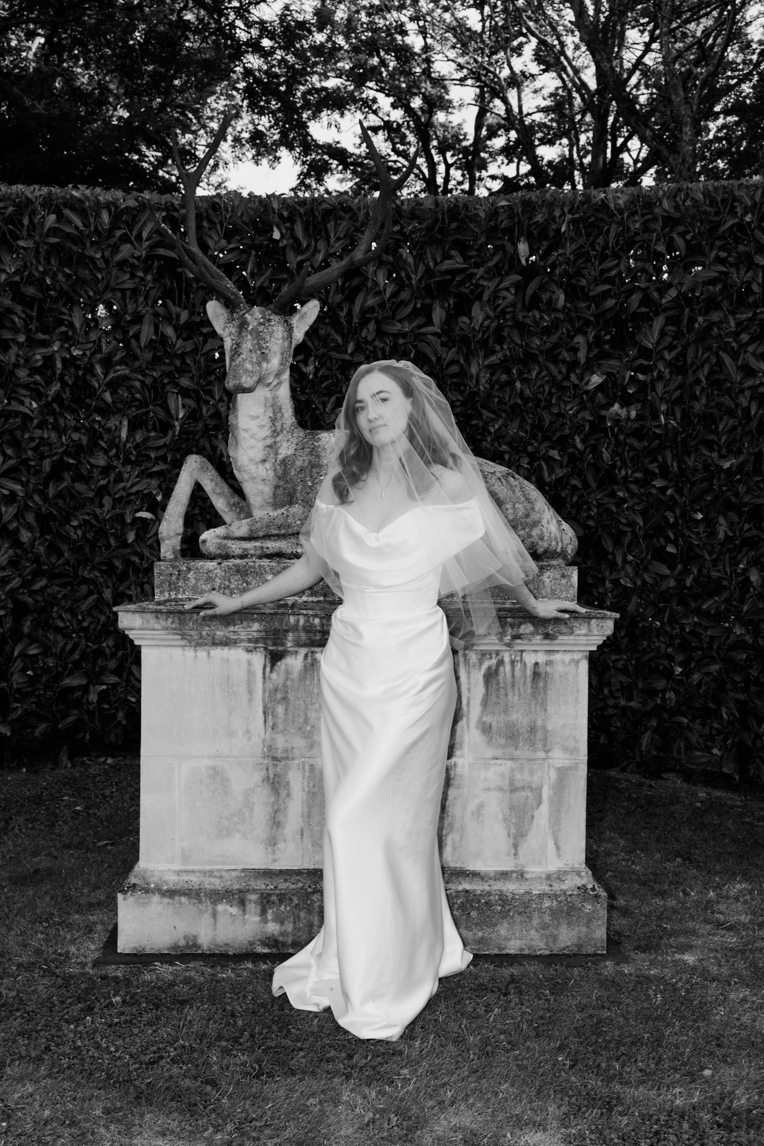 A figure in a flowing white dress poses gracefully on stone pillars in a garden setting at dusk. Luxury wedding france
