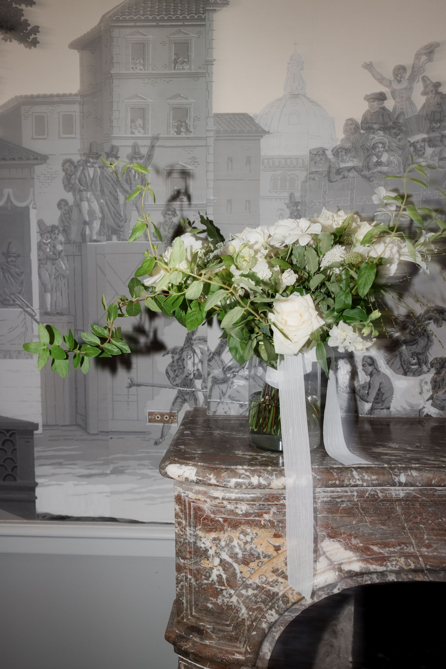 White floral arrangement with lush greenery in a tall vase against a black and white vintage photograph backdrop.
