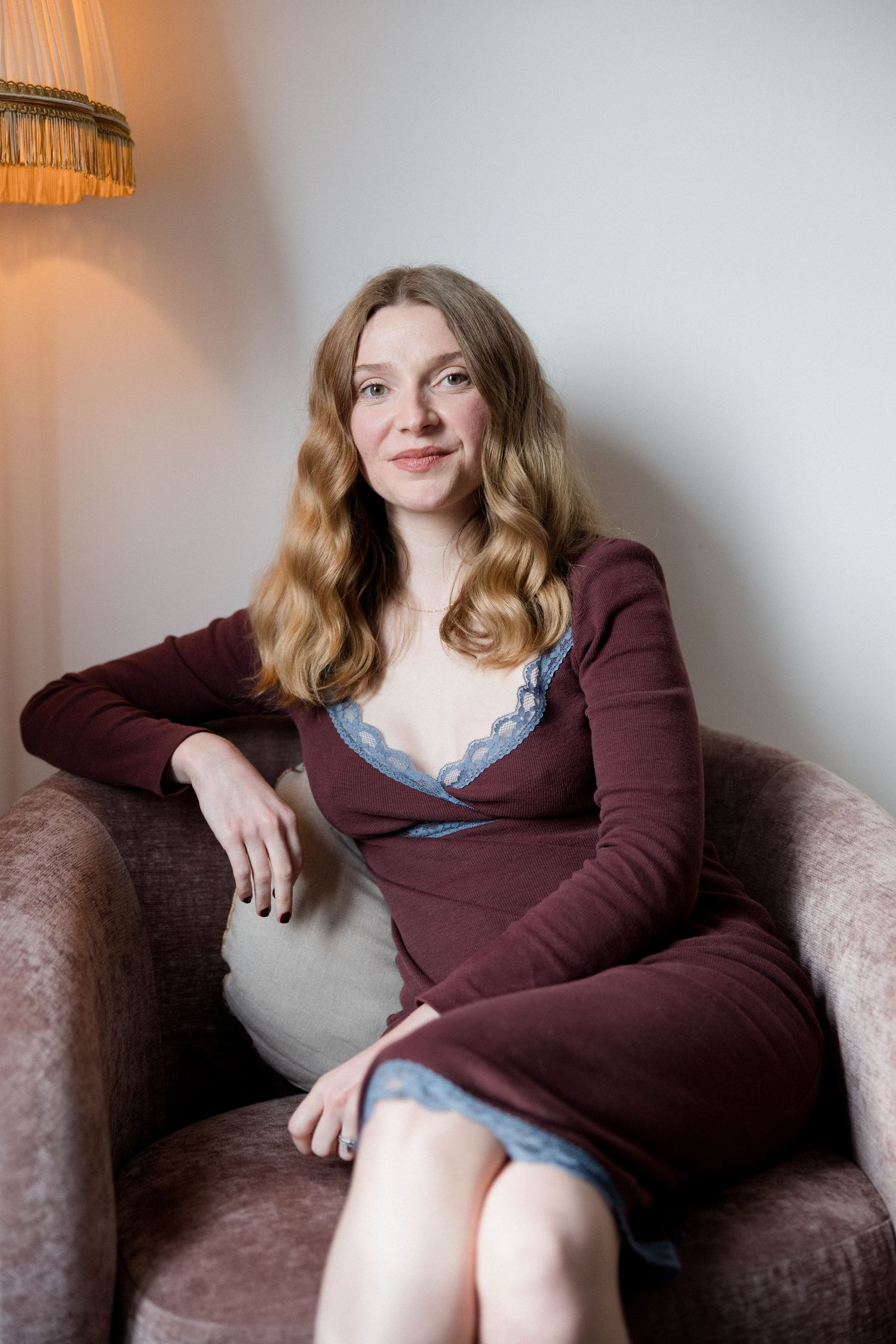 Young woman with wavy auburn hair sits relaxed in a velvet armchair, wearing a burgundy dress with lace trim.