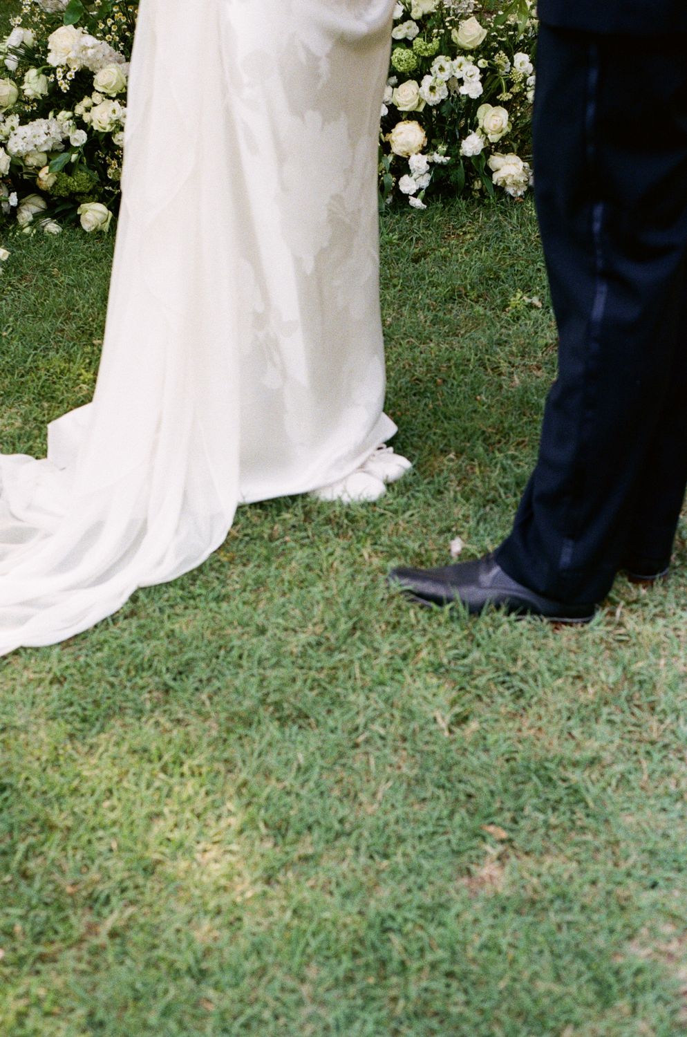 A view of black dress shoes and the hem of a white wedding dress against green grass with white flowers in the background.