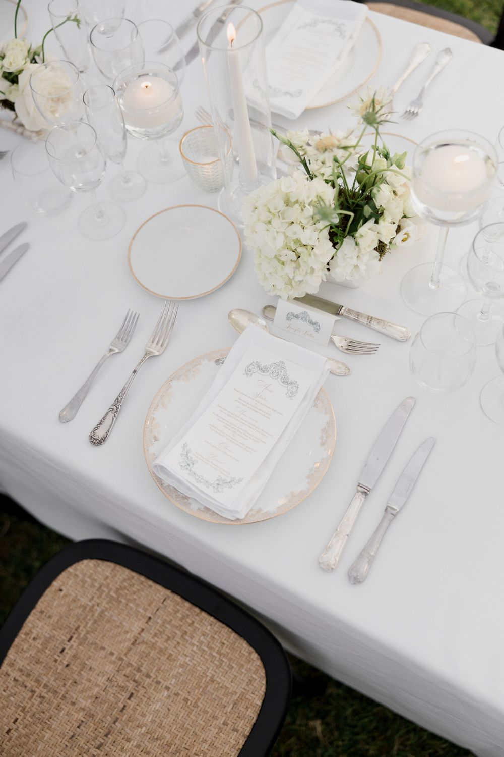 Elegant white table setting with gold-rimmed plates, silverware and soft floral centerpieces on crisp white linens.