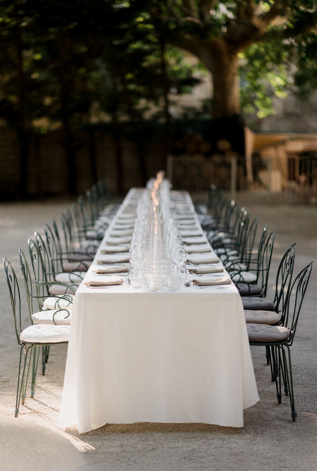 Long white banquet table with glass place settings and metal bistro chairs arranged in a shaded outdoor setting.