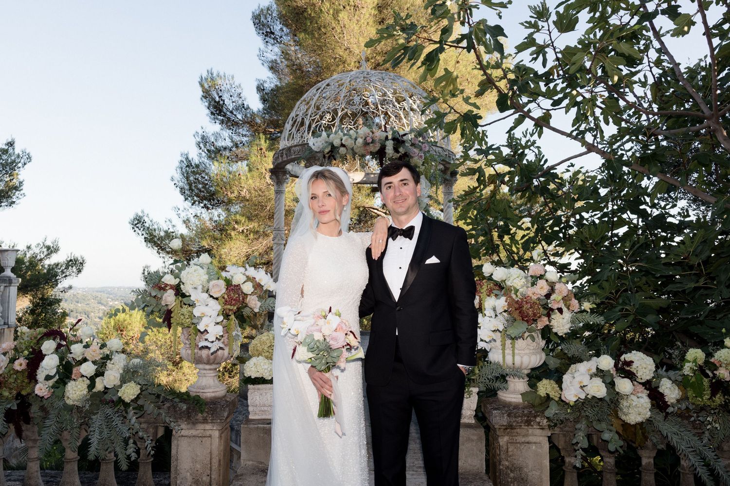 Elegant wedding couple poses under ornate floral arch with white roses and greenery in outdoor garden setting. Photographe mariage france