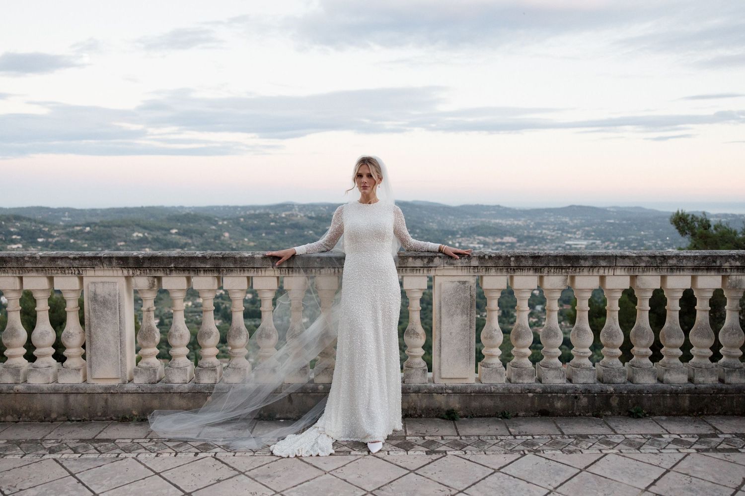 A bride in a flowing white gown stands at a stone balcony overlooking a scenic mountain landscape at sunset.