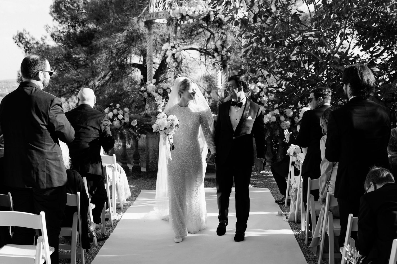 Black and white wedding party photo sequence showing guests and couple walking outdoors under trees.