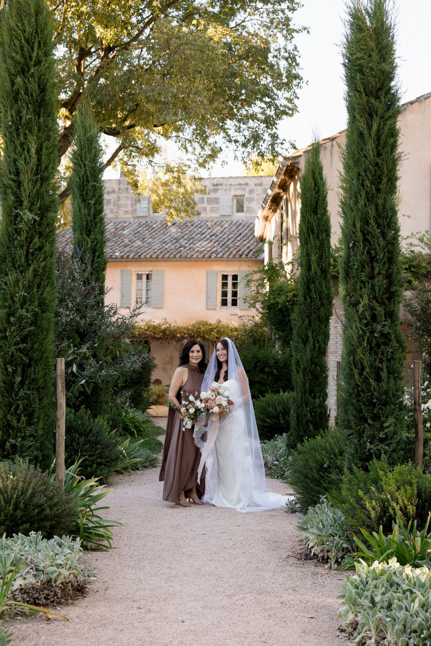 Wedding couple poses along a Mediterranean garden path lined with tall cypress trees and lush greenery.