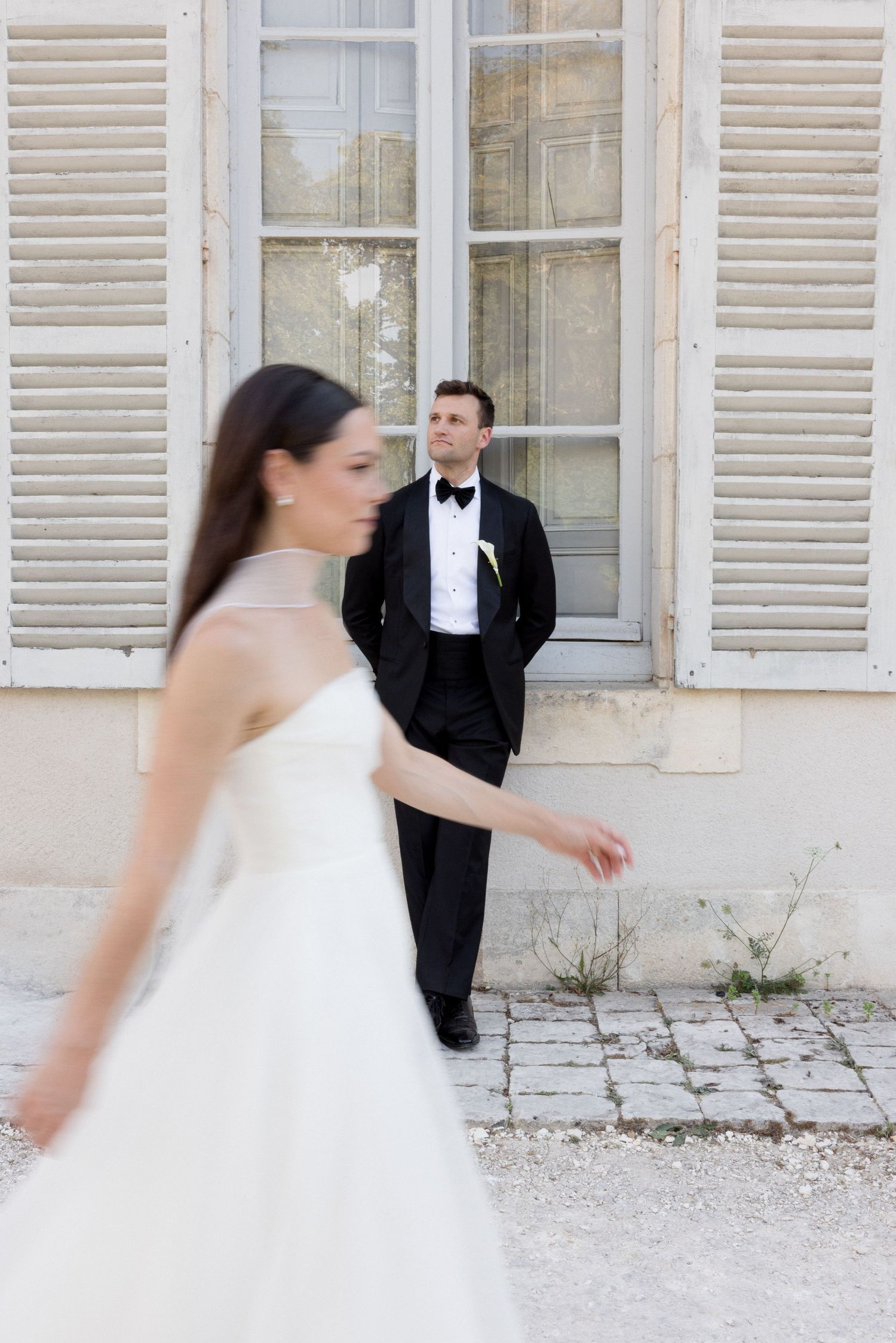 Bride in white strapless gown walks as groom in black tuxedo watches outside a French chateau.