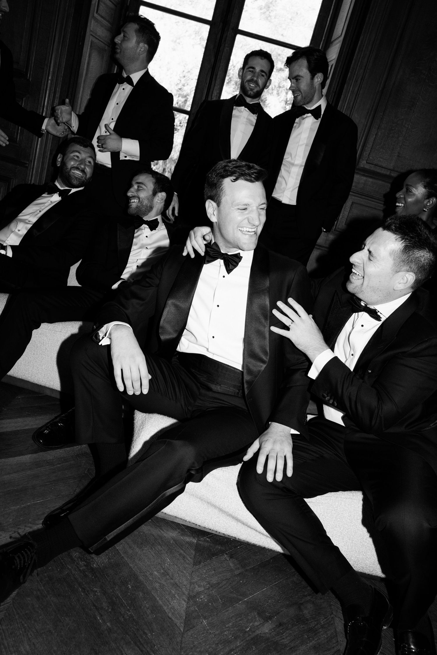 Black and white photo of groomsmen in tuxedos laughing and relaxing together at a wedding celebration.