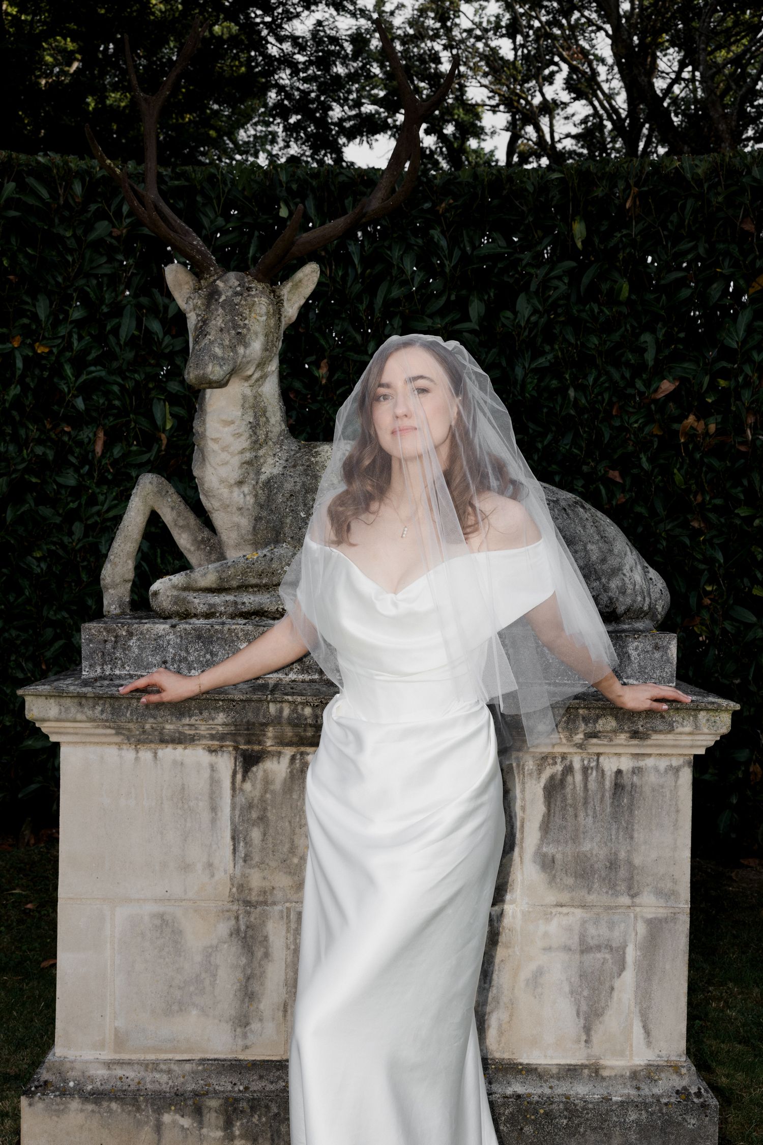 A bride in a white wedding dress and veil poses playfully in front of stone garden statues. Luxury wedding france