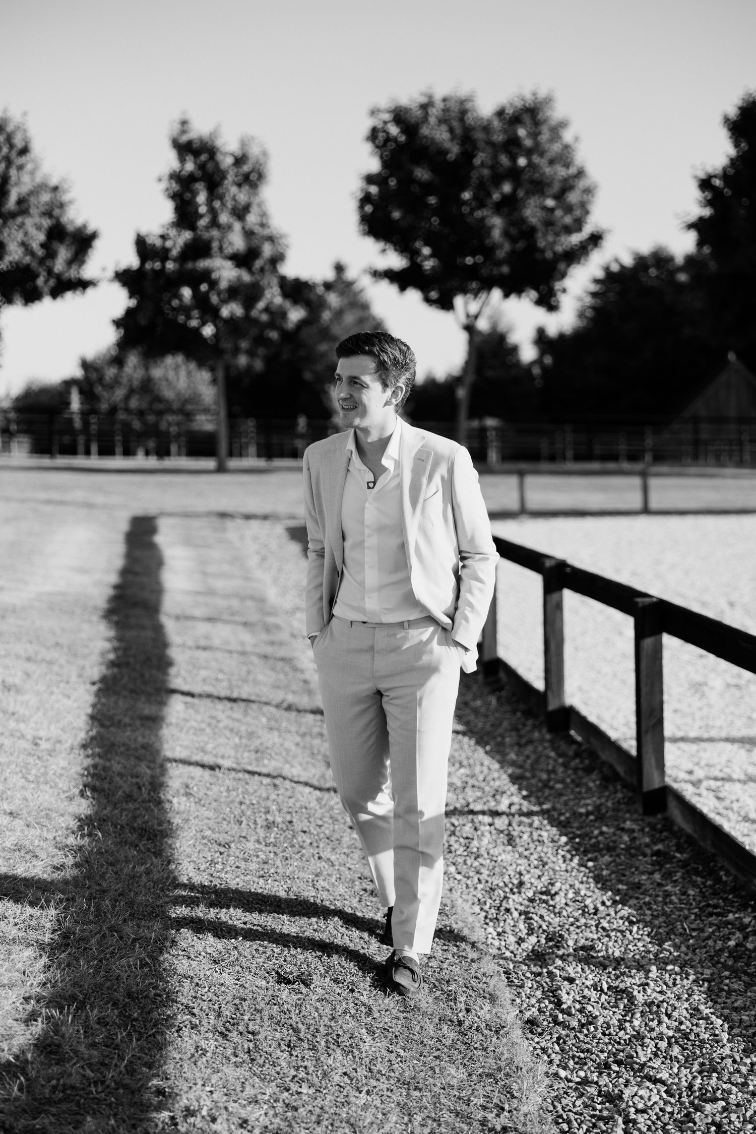 A person in white clothing walks along a fence line casting long shadows in black and white photography.
