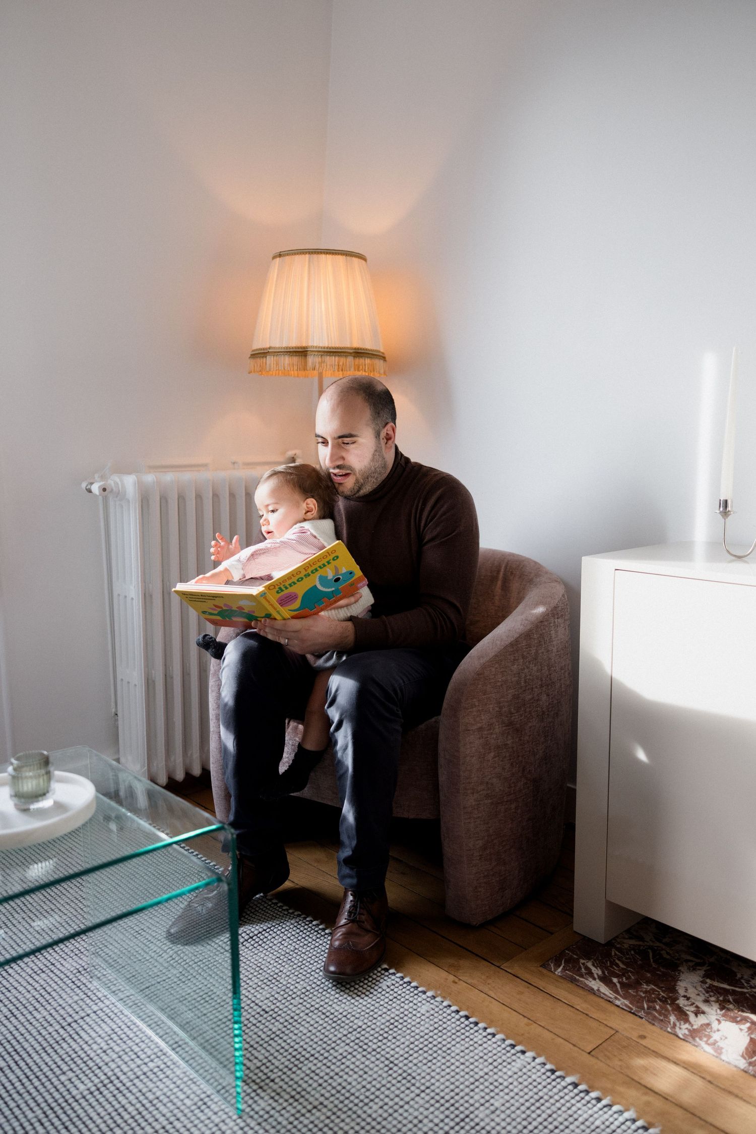 Father reads colorful picture book to toddler on armchair beside glowing lamp in cozy living room.