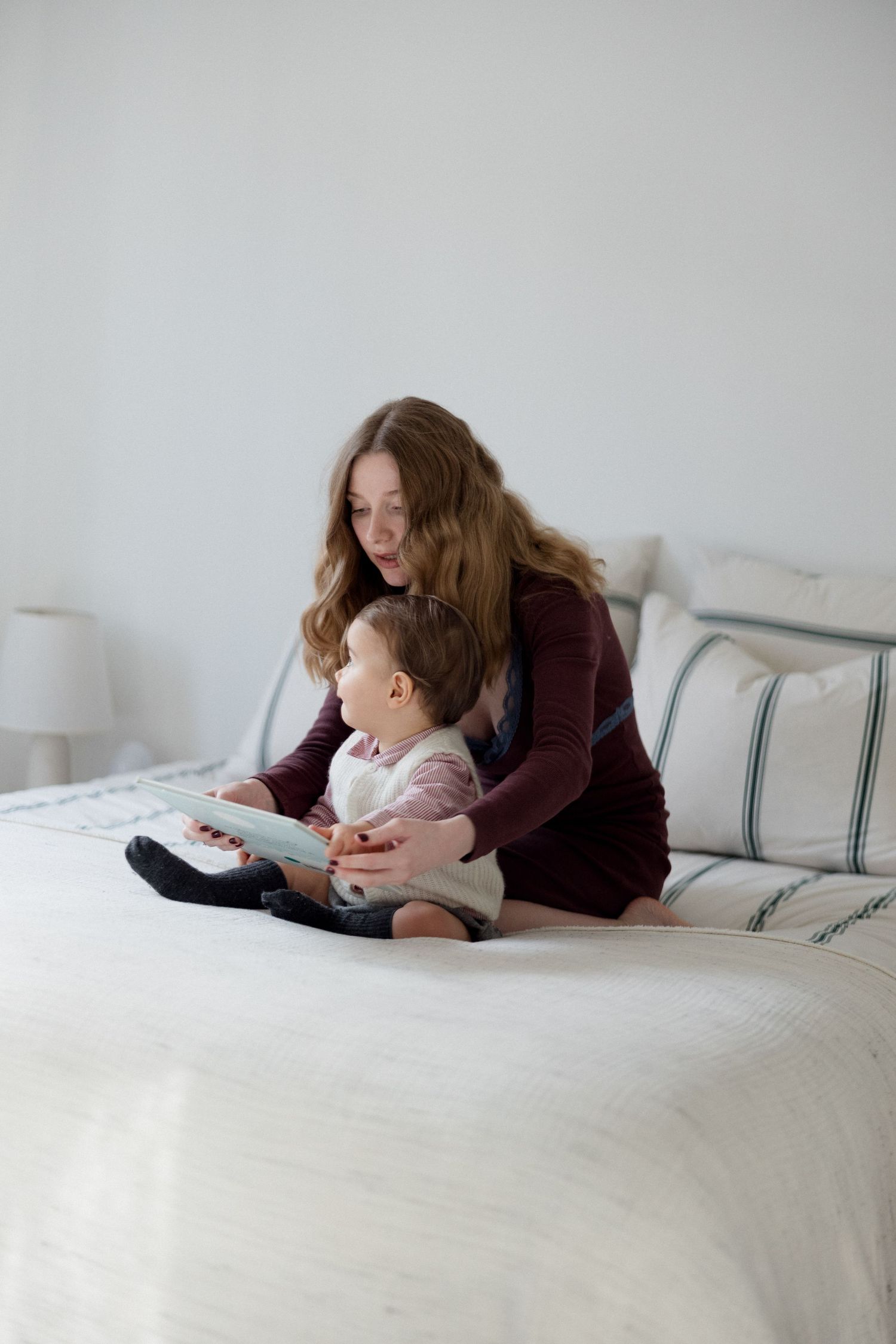 Mother sitting on bed reading a book with her toddler baby in a bright white bedroom.