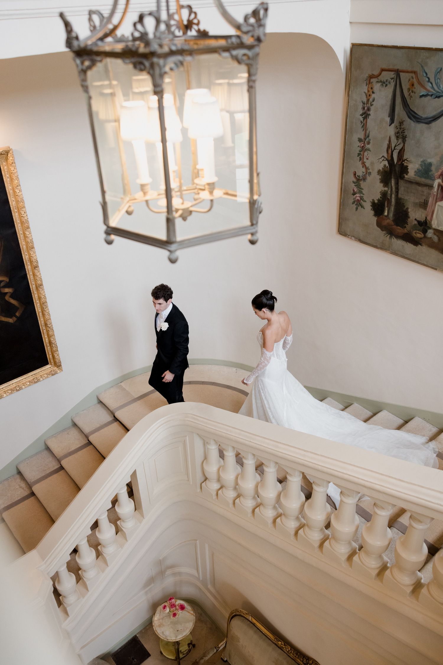 Bride and groom descend elegant curved staircase beneath ornate chandelier in grand estate wedding venue.