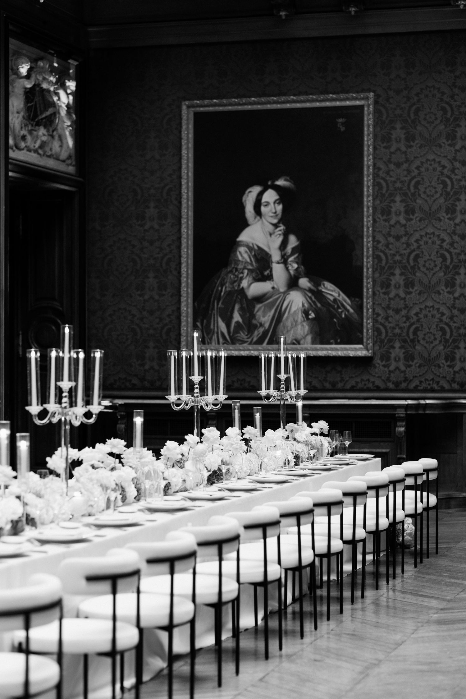 Elegant black and white photo of a formal dining table set beneath a classical portrait in an ornate room.