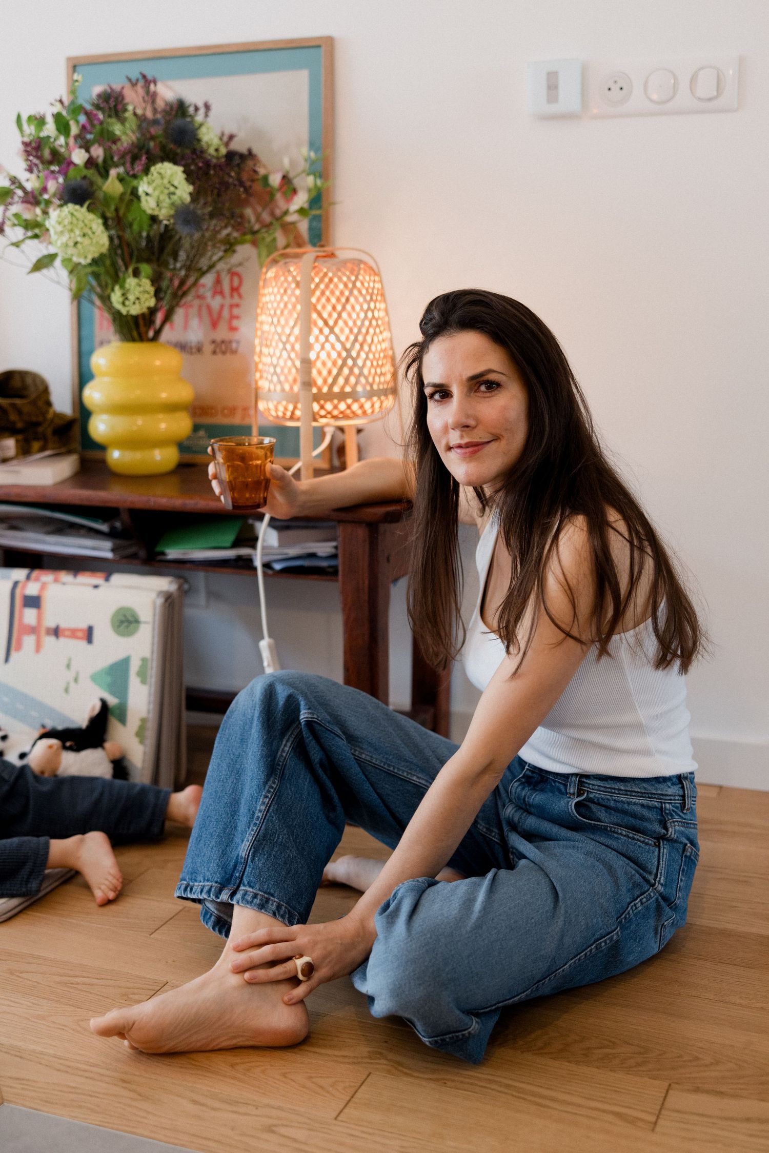 Woman in white top and jeans sits on floor holding tea beside a lamp and floral arrangement.