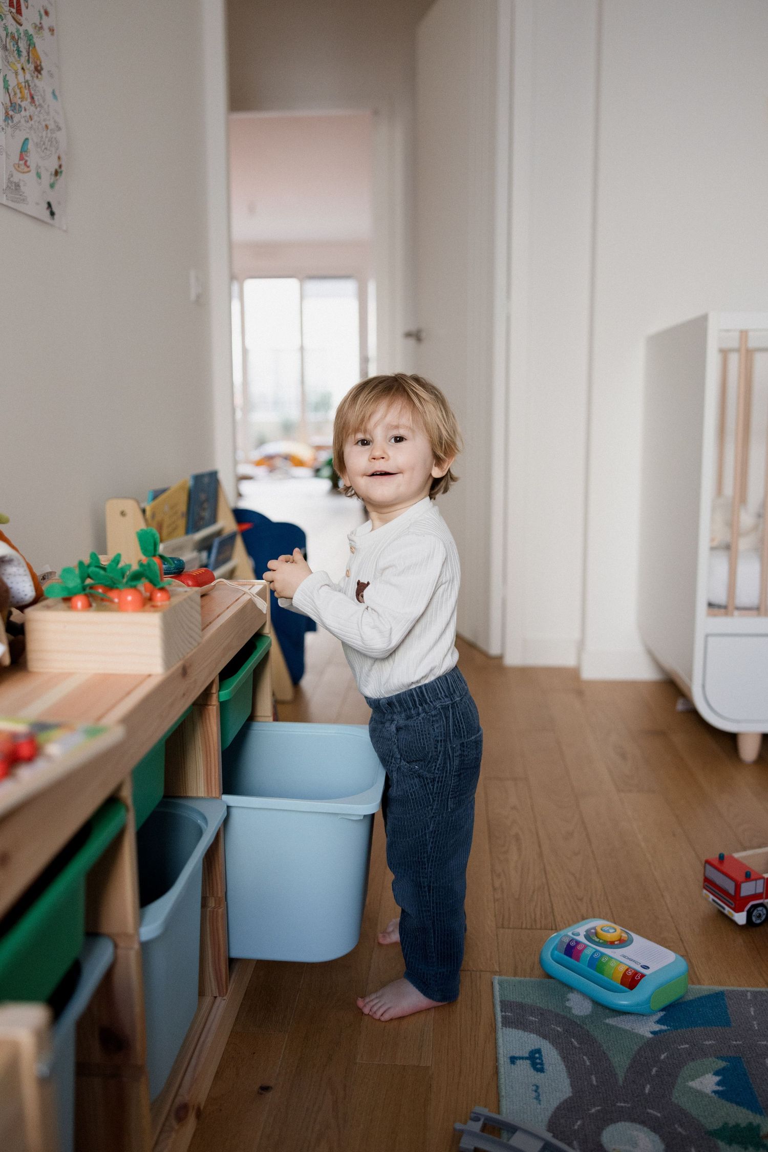 Toddler boy playing with a blue storage bin in a bright, toy-filled children's bedroom with wooden floors.