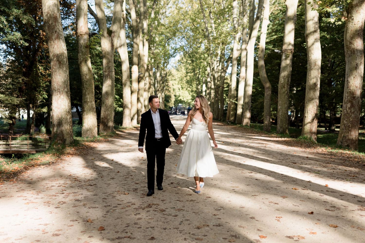 Bride and groom walk hand-in-hand along a tree-lined path in a sunlit park on their wedding day.
