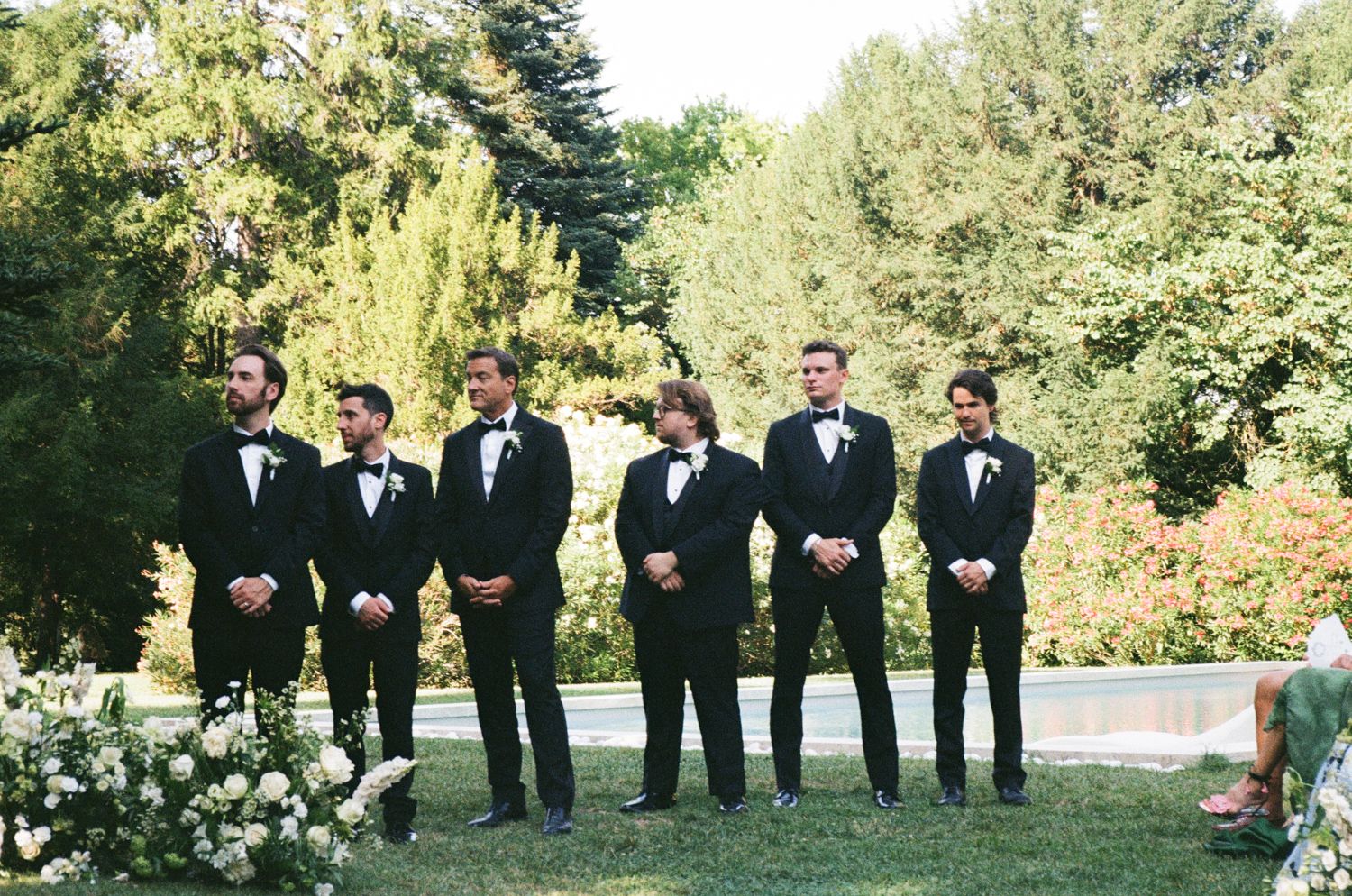 Wedding party in black tuxedos stands together on green lawn beside white rose garden during outdoor ceremony.