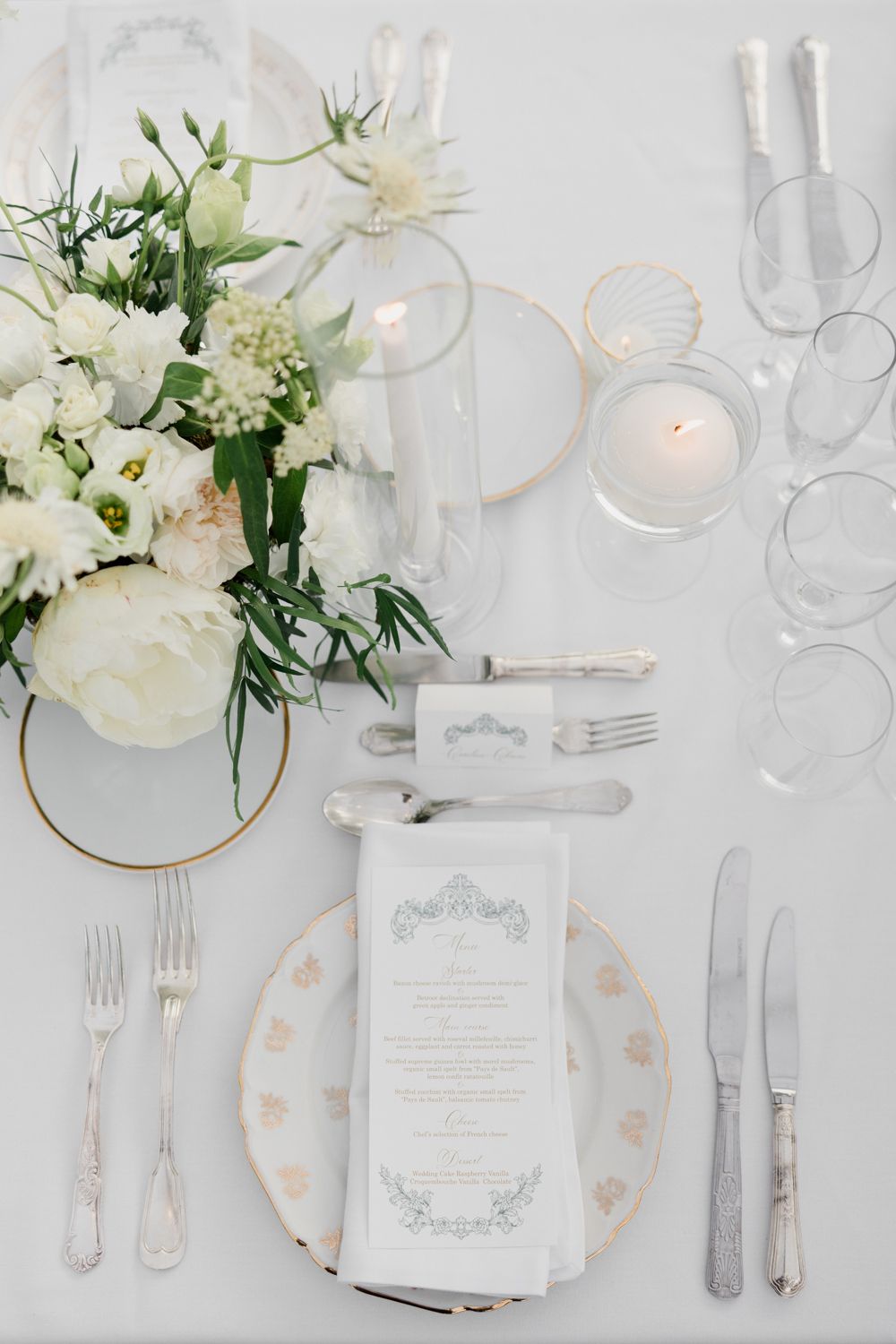 Elegant white wedding table setting with floral centerpieces, gold accents, menu cards and glowing candlelight. Château des 3 Fontaines wedd