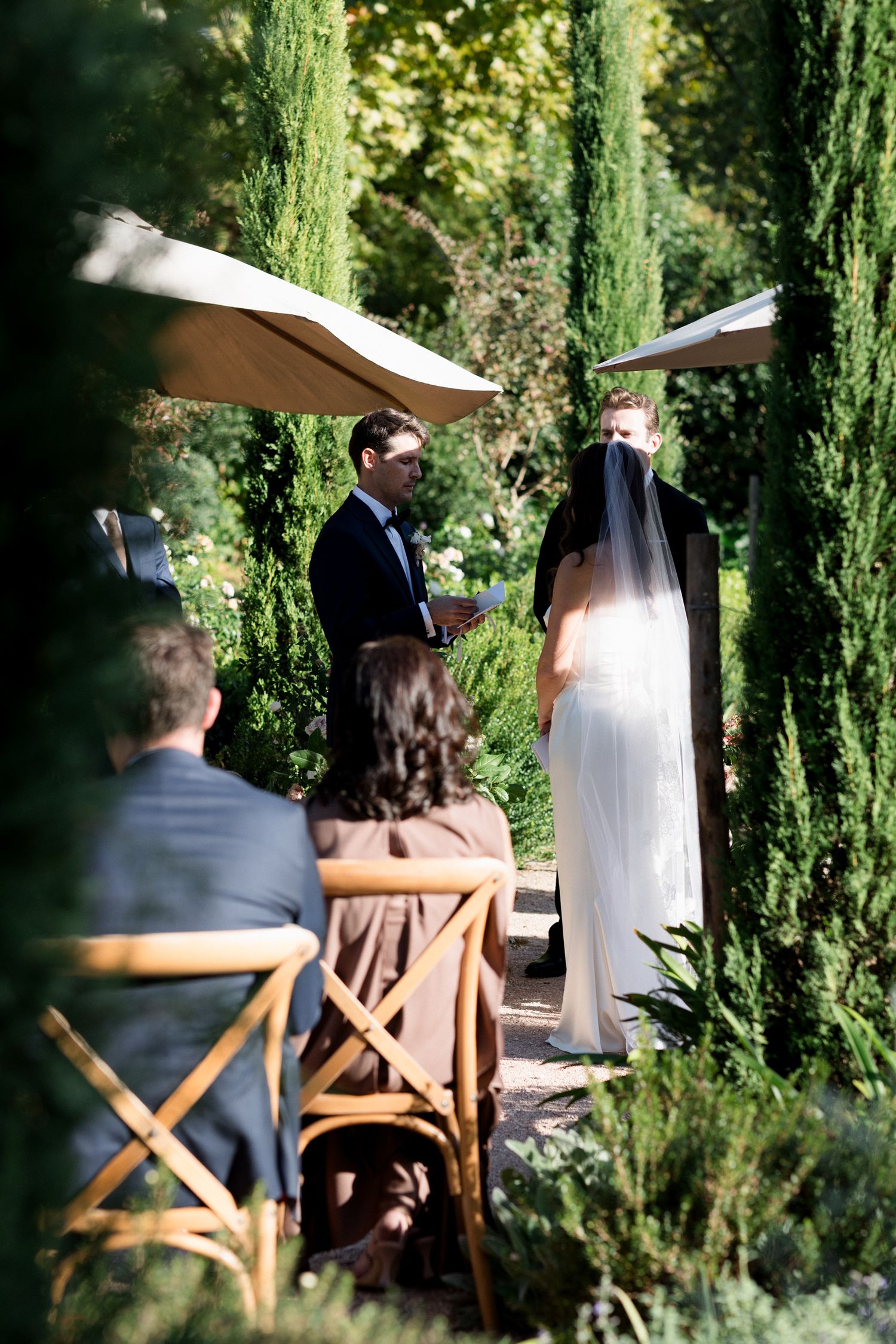 Wedding ceremony guests seated among tall cypress trees in an outdoor garden setting during a sunny day.