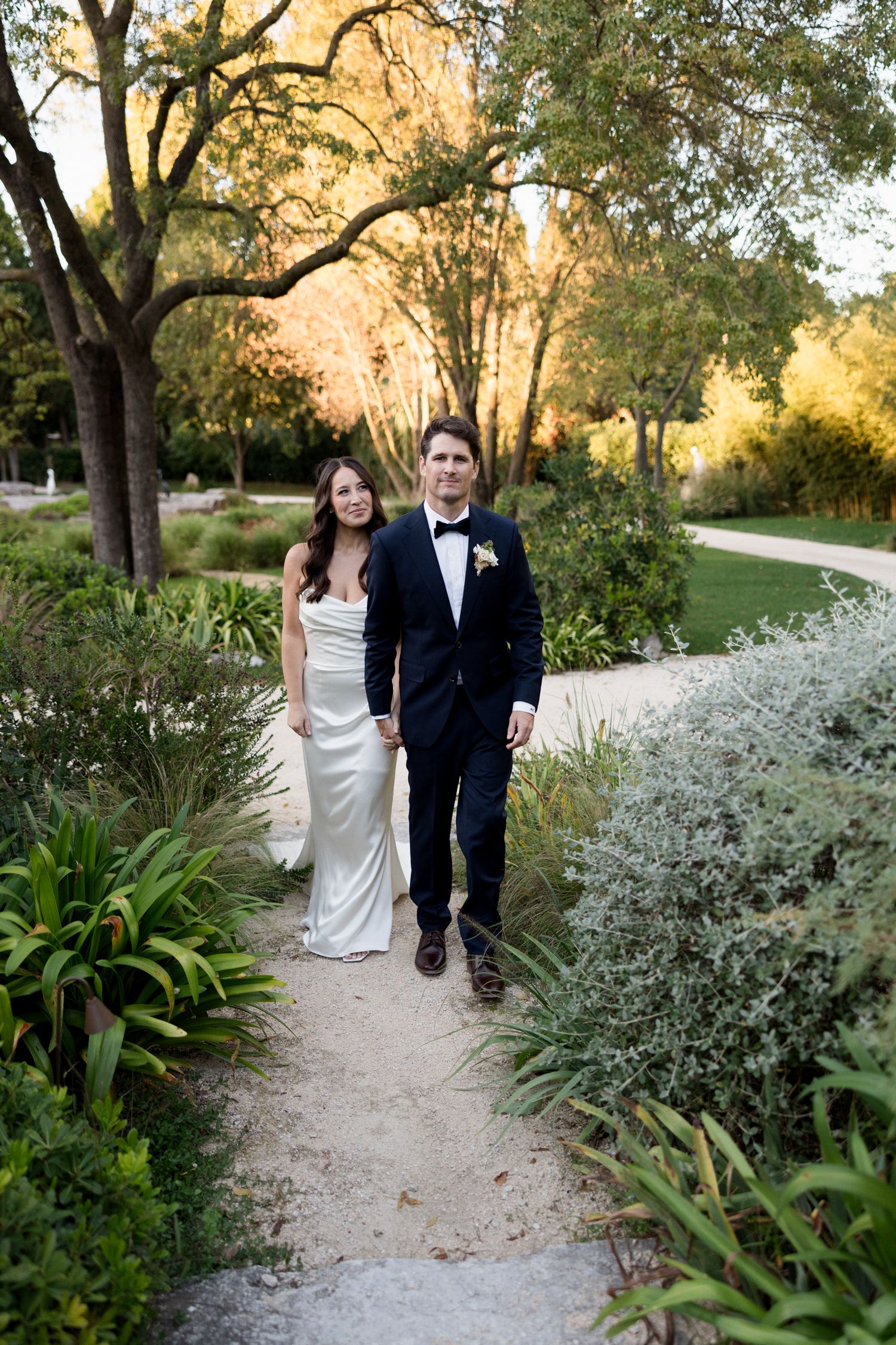 Newlywed couple walks along a garden path surrounded by white flowers and lush greenery at sunset.
